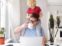 Mann im Homeoffice vor dem Laptop mit Kind auf den Schultern